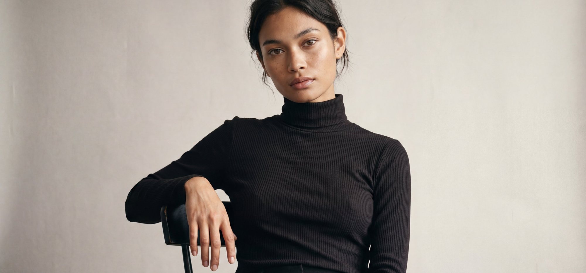 AI-generated image of a model in a black ribbed turtleneck sitting on a black chair against a light grey wall